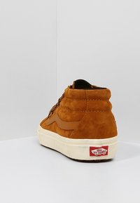 Brązowe zamszowe sneakersy wysokie z kontrastowym skórzanym paskiem po boku, teksturowana cholewka, biała gumowa podeszwa i czerwone logo Vans na pięcie.