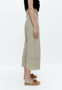 Béžové kalhoty s širokými nohavicemi a roztrhaným lemem, vyrobené z texturované tkaniny. V kombinaci s hnědými skládacími sandály s dekorativním zlatým akcentem.