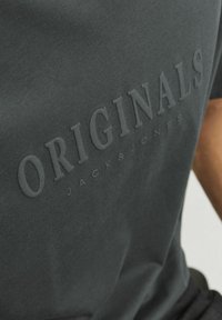 Tumši pelēkas kokvilnas T-krekls ar reljefu tekstu "ORIGINALS JACK&JONES." Īsas piedurknes un apaļa kakla izgriezums ar gludu tekstūru.