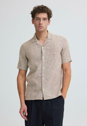 Junger Mann mit lockigem Haar, der ein beigefarbenes kurzärmeliges Hemd mit Knöpfen und dunkle Hosen trägt, steht mit einer Hand in der Tasche vor einem schlichten Hintergrund.