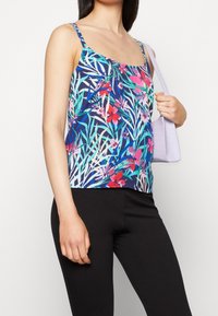 Femme portant un haut sans manches à fleurs bleu et un pantalon noir, tenant un petit sac à main violet clair sur son épaule.