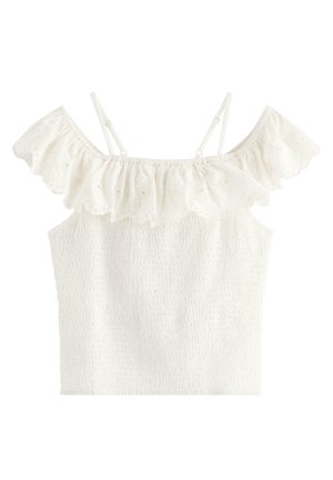 Ung pige iført hvid skulderfri top og denimshorts, der holder en hvid taske med flæser, iført beige ballerinaer med blomster.