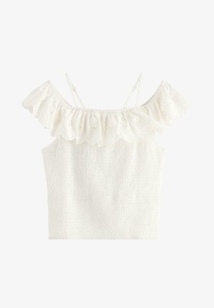 Młoda dziewczyna w białej bluzce odsłaniającej ramiona i dżinsowych szortach, trzymająca białą torebkę z falbanami, ubrana w beżowe baleriny w kwiaty.