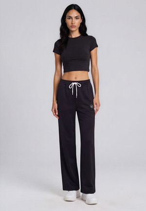 Femme debout portant un t-shirt court noir, un pantalon large noir à cordon et des baskets blanches sur un fond uni gris clair.