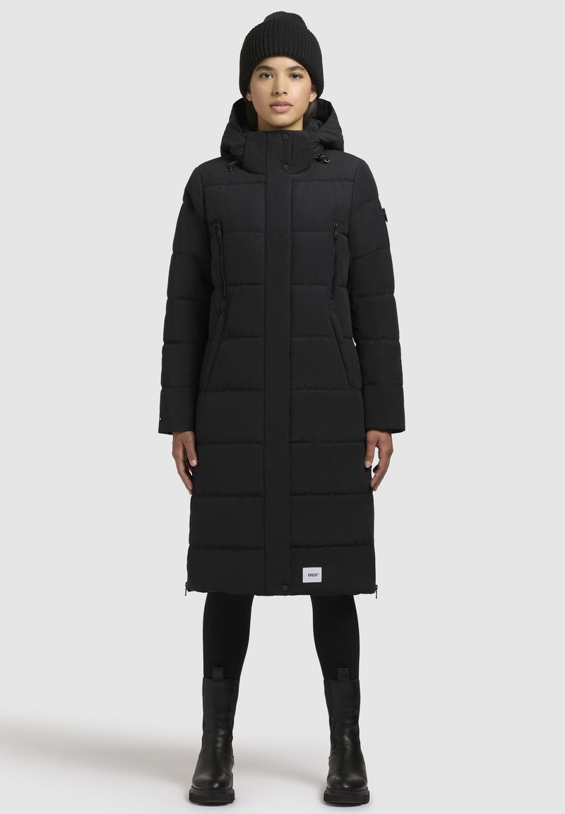 Cappotto lungo nero in piumino con colletto alto, tasche con zip e design trapuntato. Abbinato a un berretto a maglia nero e stivaletti alla caviglia.