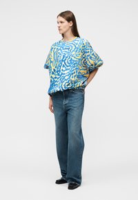 Frau trägt locker sitzendes T-Shirt mit blau-gelbem abstraktem Muster, weite blaue Jeans und schwarze Schuhe, steht mit hinter dem Rücken verschränkten Händen.