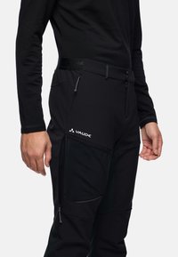 Zwarte softshell broek van gemiddelde gewicht met een zachte textuur, ritszakken en een elastische tailleband, met het "VAUDE" logo aan de voorkant.