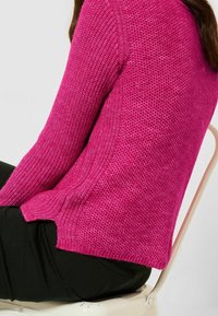 Jasnoróżowy sweter z dzianiny o fakturowanym wzorze, z bocznymi rozcięciami i ściągaczami na mankietach. Połączony z czarnymi spodniami, siedzący na jasnym fotelu.