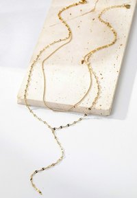 Goldene Halskette mit zwei unterschiedlichen Ketten; eine ist perlenartig, die andere im Gliederstil, beide liegen auf einer strukturierten cremefarbenen Steinoberfläche.