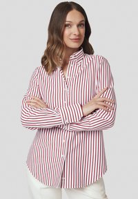 Kobieta z falującymi brązowymi włosami nosi białą koszulę z guzikami w pionowe czerwone paski, stoi z rękami skrzyżowanymi na tle jednolitym.