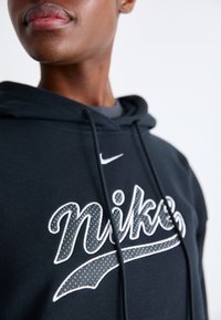 Svart hoodie med framträdande vitt Nike-logotyp och söm detaljer. Materialet är mjukt med ett texturerat mönster på logotypen. Dragsnören ingår.