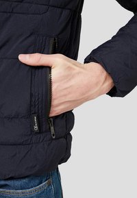 Hand teilweise in der mit Reißverschluss versehenen Tasche einer schwarzen gesteppte Jacke, die über blauen Jeans getragen wird, vor einem hellen Hintergrund.