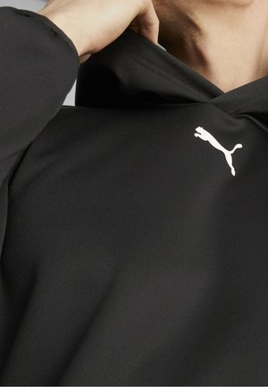 Persoon draagt een zwart Puma-hoodie, trekt met de hand de kraag omhoog, toont het witte Puma-logo op de bovenborst.