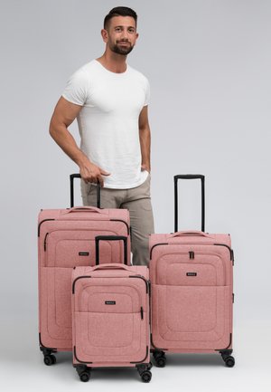 Trois valises roses de tailles variées avec un tissu texturé, des accents noirs et des poignées rétractables, présentées aux côtés d'un homme debout.