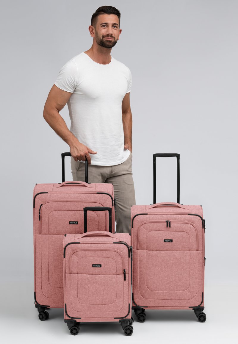 Drie roze koffers in verschillende maten met een gestructureerde stof, zwarte accenten en uitschuifbare handvatten, naast een staande man getoond.