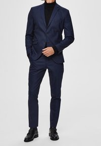 Mężczyzna w dopasowanym granatowym garniturze, czarnym golfie i czarnych butach formalnych, stoi z jedną ręką w kieszeni, a drugą poprawiając żakiet.