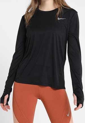 Femme portant un t-shirt noir à manches longues Nike et des leggings orange rouille avec des logos Nike, debout devant un fond blanc.