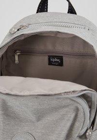 Šedý plátěný batoh uvnitř s kapsou na zip, štítkem Kipling a měkkou podšívkou. Má zaoblený klapkový uzávěr a kovové detaily.