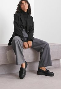 Zwarte loafer schoenen van gestructureerd leer, met dikke zolen en een kettingaccent. Draagt u met grijze wijde broek en een zwarte blazer.