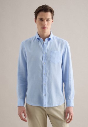 Junger Mann, der ein hellblaues, langärmliges Hemd mit Knöpfen und beige Hose trägt, steht vor einem einfarbigen Hintergrund.