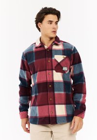 Man in een knoopjesburgundy, blauwe en crèmekleurige grote geblokte flanellen shirt met beige broek, kijkend naar links.