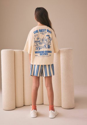 Kind staat met de rug naar de kijker, draagt een beige oversized t-shirt met blauwe brunch cartoonprint en gestreepte korte broek tegen een beige achtergrond.