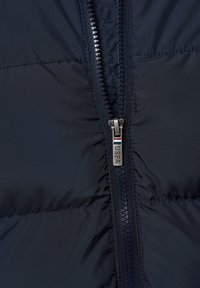 Navy pufferjack stof met een gequiltde textuur, voorzien van een stevige ritssluiting aan de voorkant en een zilveren U.S. Polo Assn. logo op de ritssluiting.
