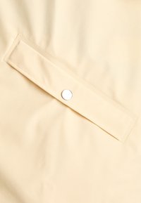 Țesătură bej, netedă, cu un finisaj lucios; prezintă o clapetă cu un buton de prindere argintie. Design minimalist, fără modele vizibile.