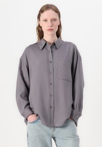 Camisa cinza de corte relaxado, de mangas longas, com finas riscas verticais em rosa, fechamento com botões na frente e um bolso de remendo do lado esquerdo.