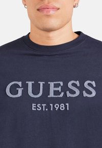 Camiseta de algodón de cuello redondo en azul marino con el logotipo bordado en gris "GUESS" y el detalle "EST. 1981" en la parte frontal. Textura suave.