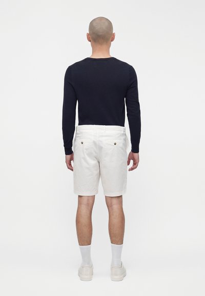 Homme à la tête rasée portant un t-shirt noir à manches longues, un short blanc jusqu'aux genoux, des chaussettes blanches et des baskets blanches, debout de dos.