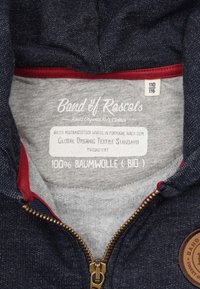 Mørkeblå bomulds-hættetrøje med en grå inderside. Har en gylden lynlås, røde detaljer og et mærke, der angiver økologisk materiale og produktionsstandarder.