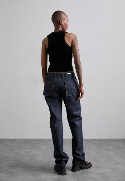 Top cu bretele negre, cu o textură canelată, asortat cu blugi largi din denim închis la culoare, prevăzuți cu buzunare mari pe spate și detalii de cusătură contrastante.