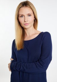 Sötétkék kötött pulóver, lekerekített nyakkivágással, hosszú ujjakkal és bordázott mandzsettával, egy hosszú, egyenes hajú személyen.