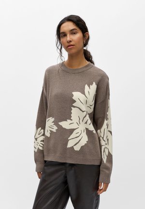Femme aux cheveux foncés portant un pull marron avec de grands motifs de feuilles blanches et un pantalon noir sur un fond blanc uni.
