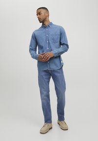 Camisa de mezclilla azul clara con un bolsillo frontal, cuello abotonado y mangas largas, combinada con pantalones de mezclilla azul a juego y zapatos beige.