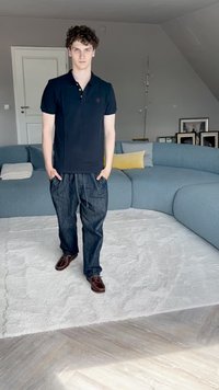 Černé polo tričko, volné tmavé džínové kalhoty s kapsami, hnědé boty. Zakřivená světle modrá pohovka a bílý plyšový koberec v minimalistickém pokoji.
