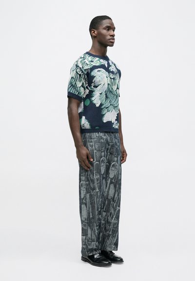 Πλεκτό πουλόβερ με πράσινο λουλουδάτο σχέδιο σε σκούρο μπλε φόντο, συνδυασμένο με φαρδιά, υφασμένα γκρι παντελόνια με αφηρημένα σχέδια. Μαύρα παπούτσια.