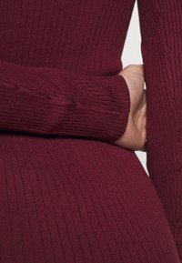 Detailní záběr na proužkovaný vínový svetr s dlouhými rukávy a texturovanou tkaninou. Materiál má hladkou, elastickou vlastnost s jemnými odlišnostmi.