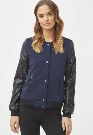 Chaqueta bomber de color azul marino con mangas de cuero negro y botones a presión. Presenta bolsillos laterales y puños y dobladillo acanalados. Textura suave.
