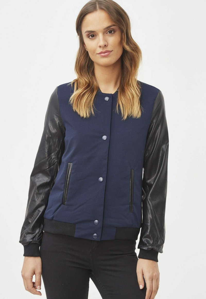 Chaqueta bomber de color azul marino con mangas de cuero negro y botones a presión. Presenta bolsillos laterales y puños y dobladillo acanalados. Textura suave.