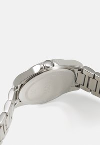Montre en acier inoxydable argenté avec un boîtier élégant, une couronne ronde et un bracelet à maillons. L'arrière présente le logo de la marque et des détails sur la résistance à l'eau.