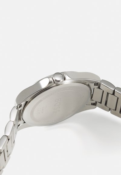 Montre en acier inoxydable argenté avec un boîtier élégant, une couronne ronde et un bracelet à maillons. L'arrière présente le logo de la marque et des détails sur la résistance à l'eau.