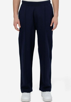 Uomo che indossa pantaloni blu navy a taglio largo con vita elasticizzata e sneakers bianche, in piedi davanti a uno sfondo neutro.