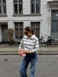 Polo majica z dolgimi rokavi z belimi, sivimi in črnimi črtami, kombinirana z modrimi širokimi kavbojkami. Drži svetlo roza torbico.