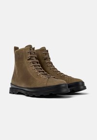 Botas de ante marrón hasta el tobillo con puntera redonda, cuello acolchado, suela plana de goma negra y cierre con cordones. Textura suave y costuras mínimas.