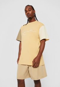 T-shirt à manches courtes en jaune clair avec des accents beiges, comprenant un logo blanc brodé. Associé à un short beige, le tout en tissu coton.