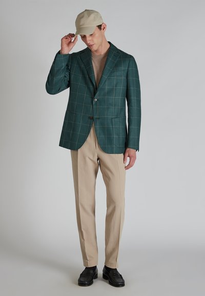 Zielona marynarka w kratę z teksturowanej tkaniny, zestawiona z beżowymi spodniami i czarnymi butami. Model nosi jasną czapkę i jasnobrązowy top.