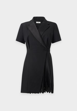 Ung kvinde stående iført en sort kortærmet blazer-kjole med plisserede detaljer, hvide sokker og sorte flade sko på en ensfarvet baggrund.