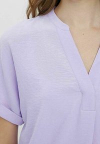 Blouse courte à manches courtes lavande avec un col en V et des manches retroussées, présentant un tissu texturé et un éclat subtil.